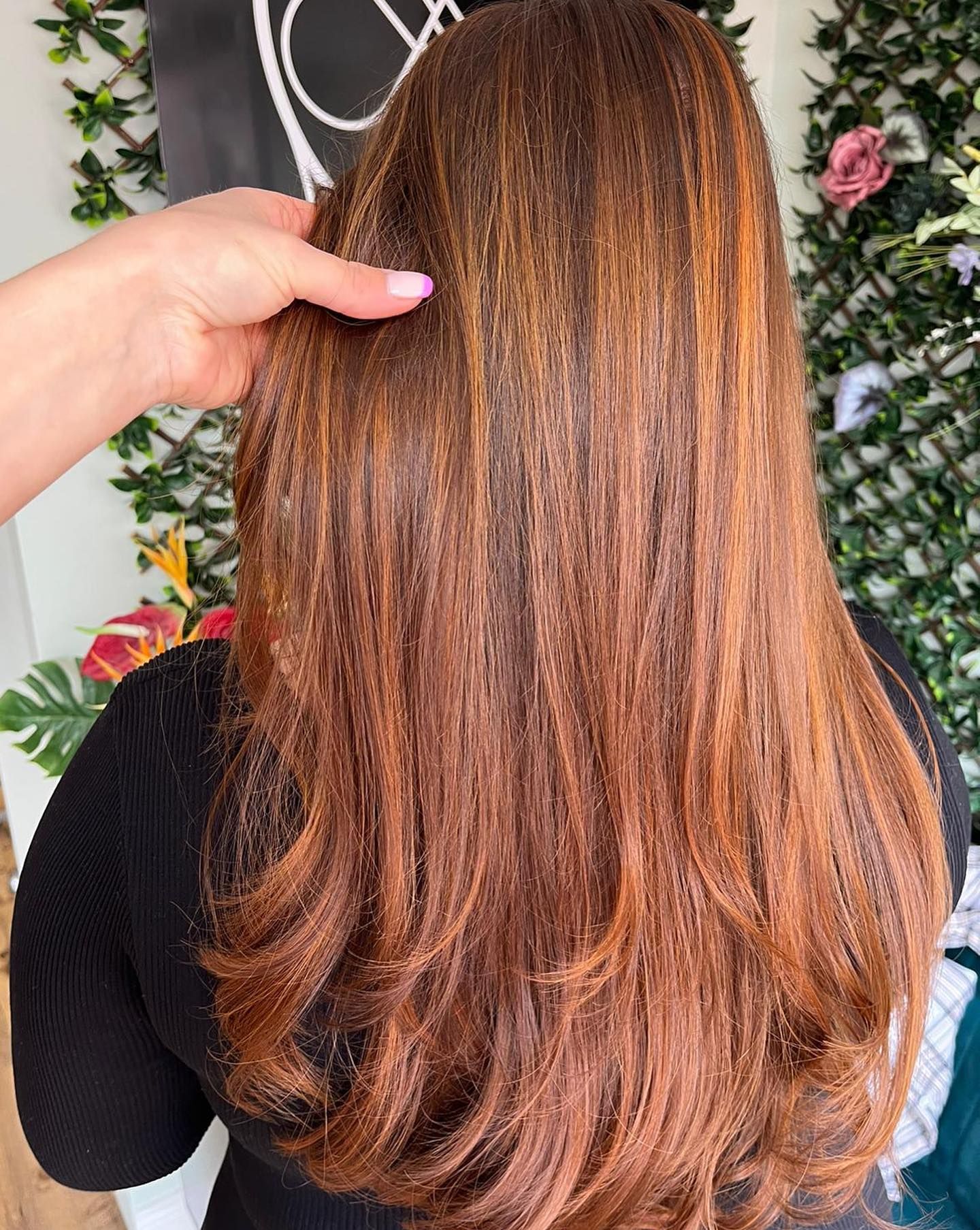 A hand gently lifts long, layered hair featuring a warm, multidimensional copper and auburn balayage.