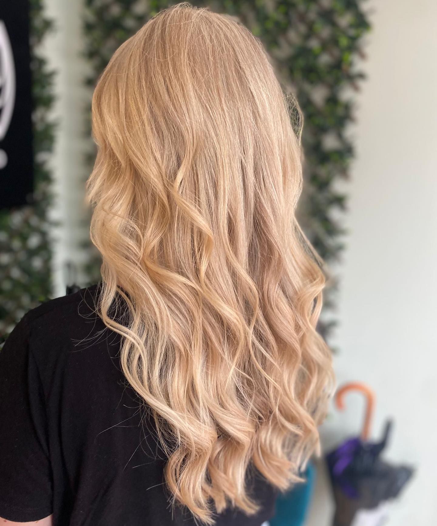 A person with long, wavy blonde hair styled in loose curls, viewed from behind against a greenery backdrop.