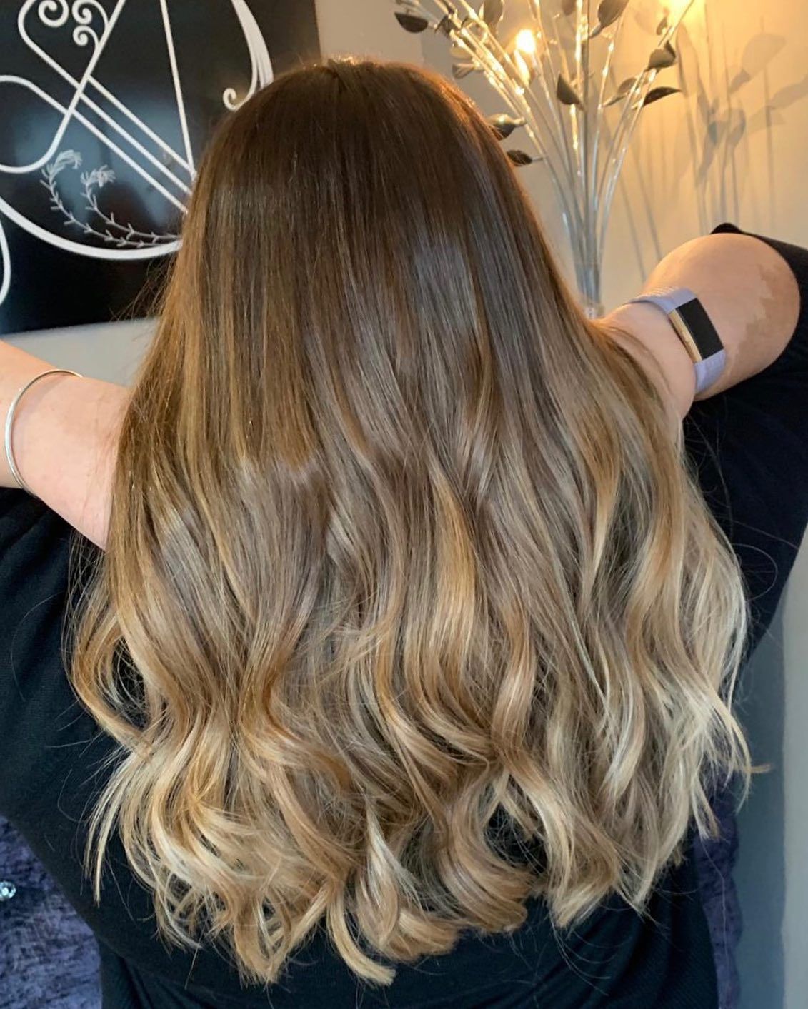 A person with wavy, brown-to-blonde ombre hair, seen from behind with hands raised to their head.