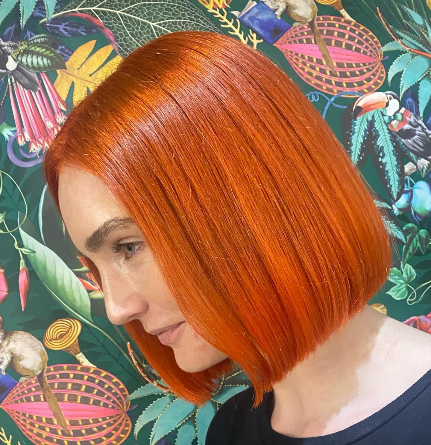 A person with a sleek, vibrant orange blunt bob haircut positioned in front of a patterned botanical wallpaper.