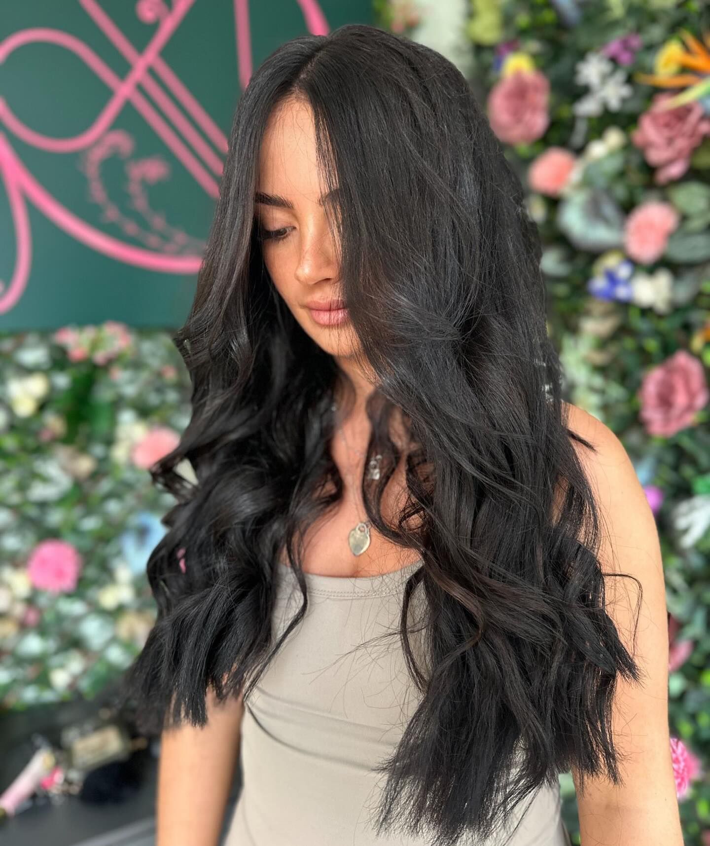 A person with long, dark, wavy hair stands in front of a decorative floral wall.