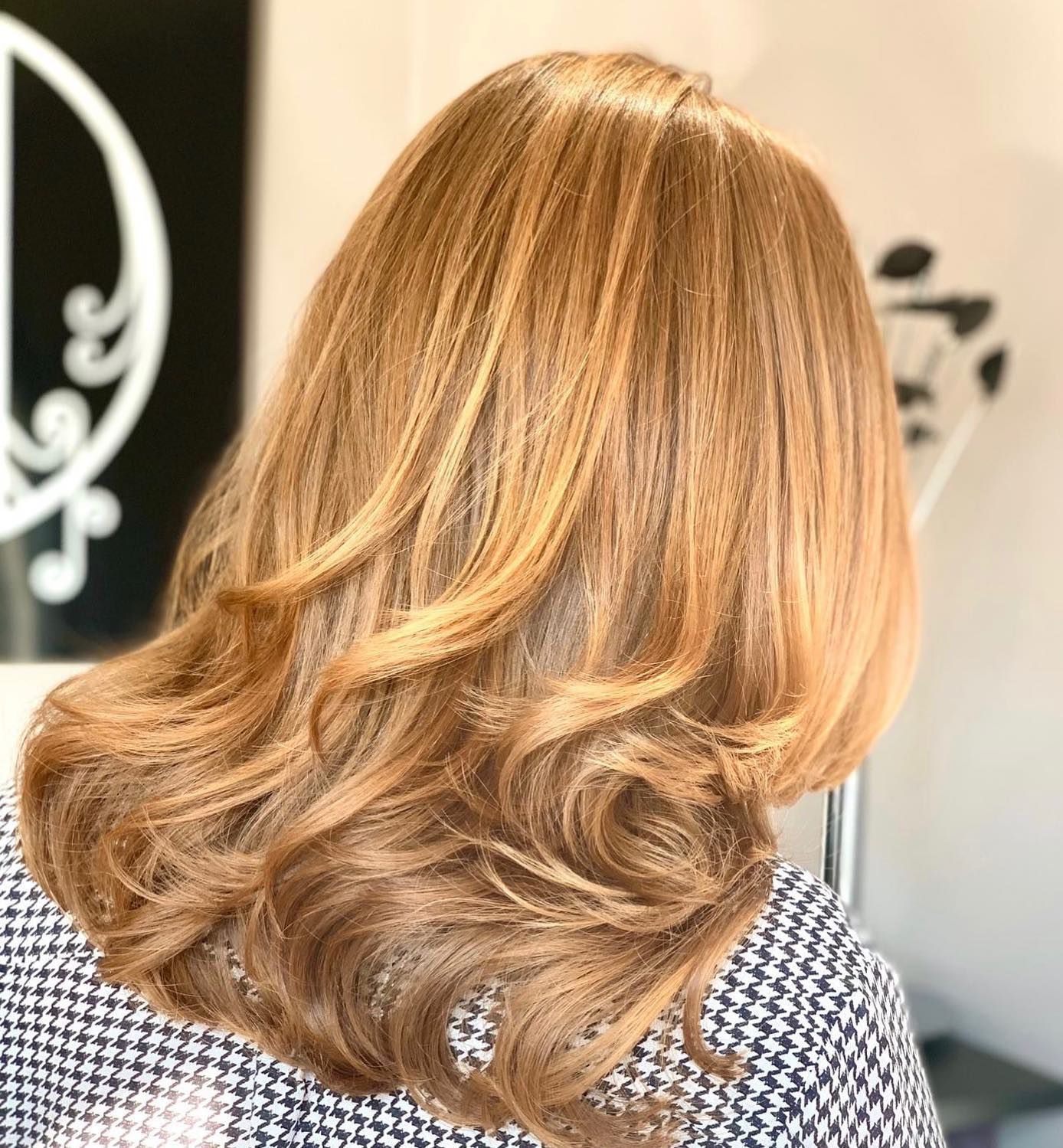 A back view of hair with soft curls and warm, honey-brown highlights, styled in a salon setting.