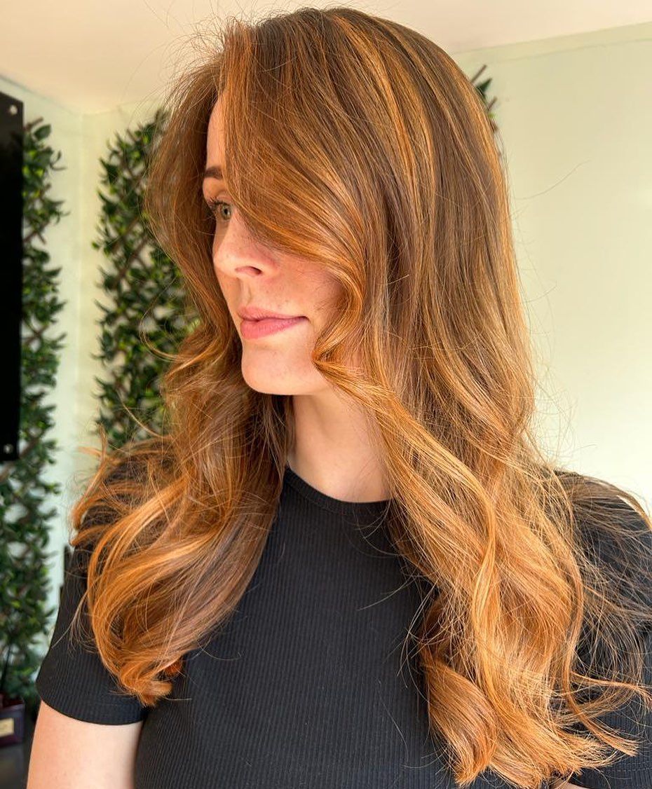 A person with long, wavy, caramel-highlighted brown hair styled with loose curls and a side-swept fringe.