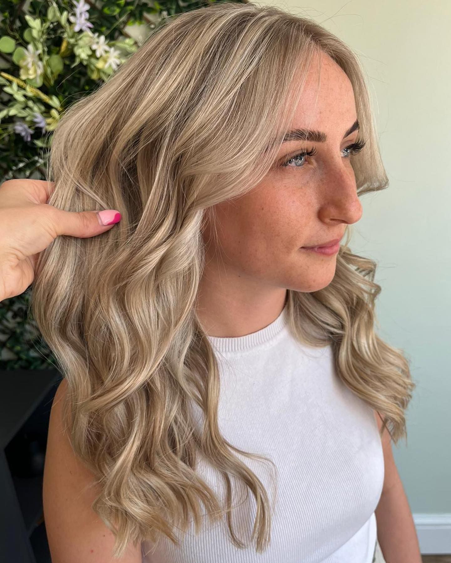 A person with wavy, blonde-highlighted hair styled with face-framing layers, shown against a floral backdrop.