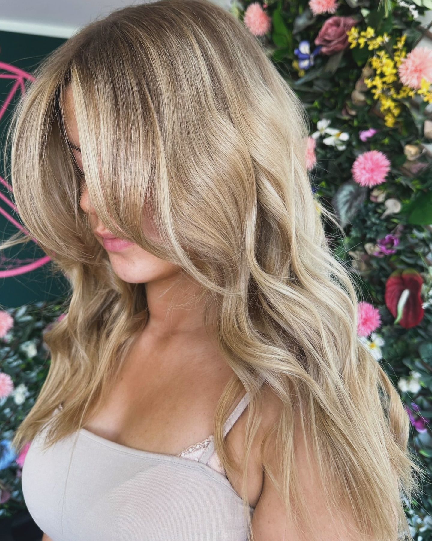 A person with long, layered, wavy blonde hair featuring face-framing bangs, standing in front of a colorful flower wall.