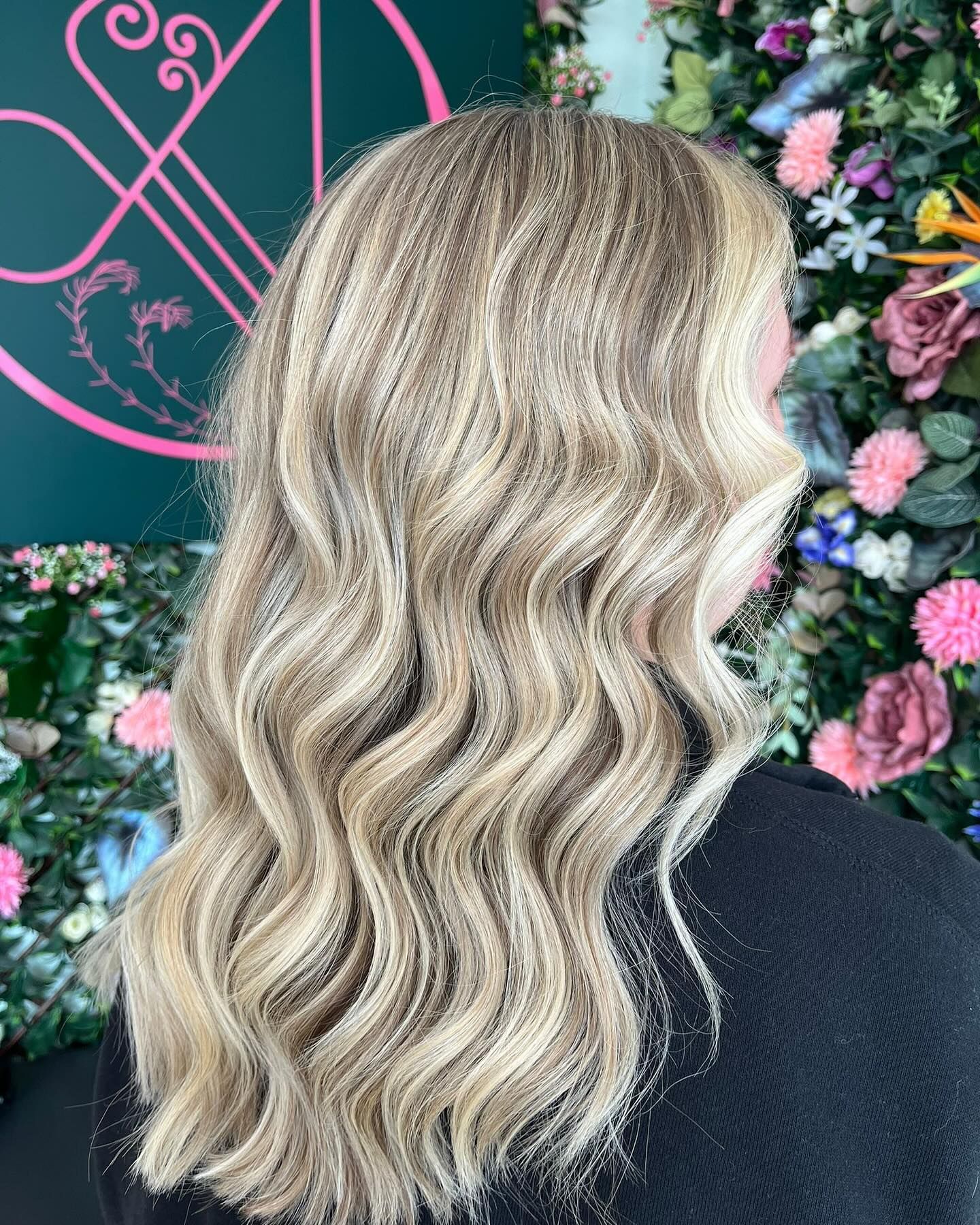 A person with highlighted, wavy, blonde hair seen from behind, positioned against a floral wall backdrop.
