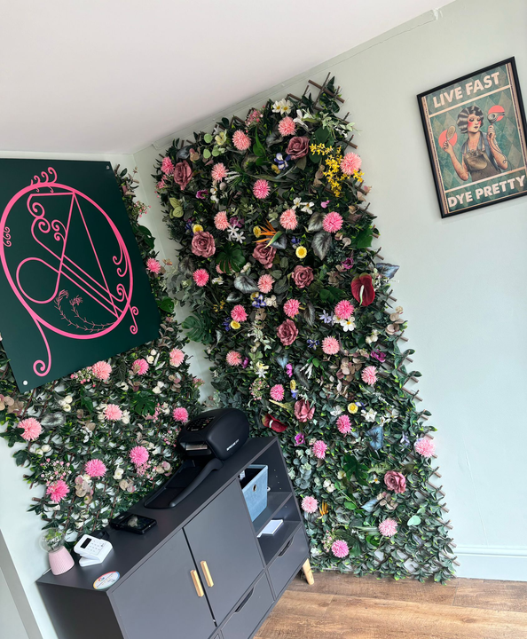 Two tall panels covered in artificial green foliage and pink roses, with a dark cabinet and a wall sign in a light room.