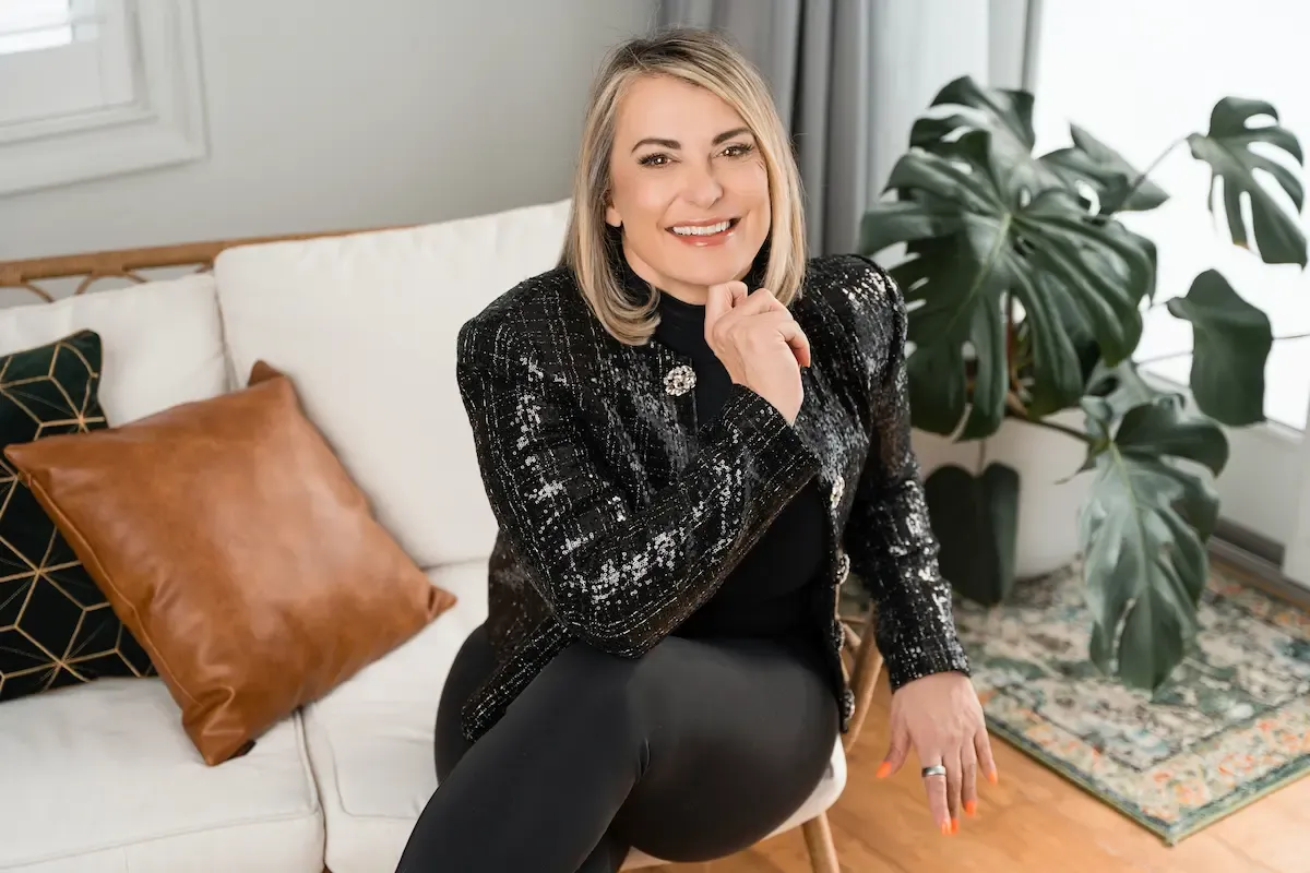 A smiling person in a sequined jacket sits on a white sofa next to a large potted plant and leather throw pillows.