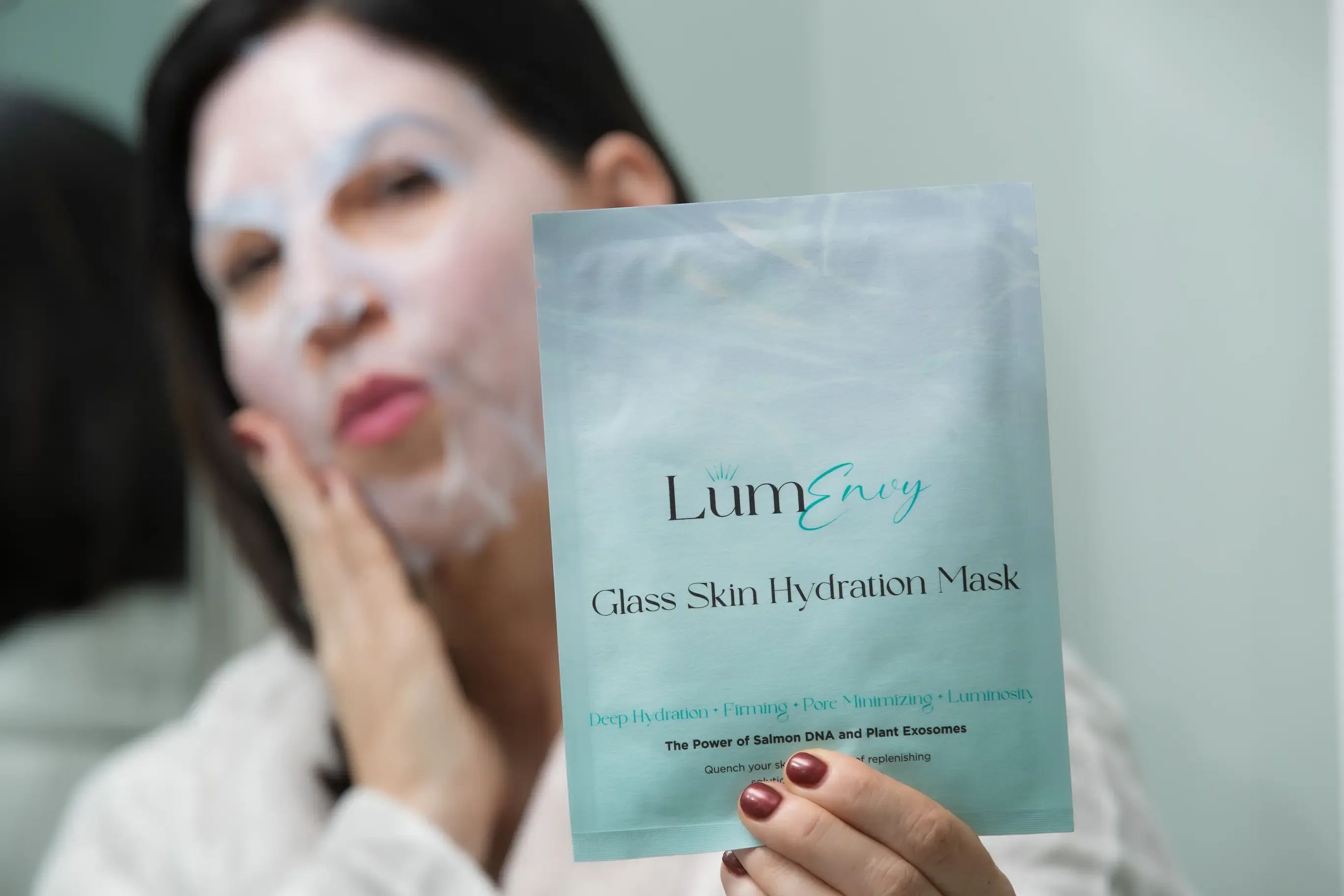 A person wears a white sheet face mask while holding a matching LumEvey Glass Skin Hydration Mask packet in the foreground.