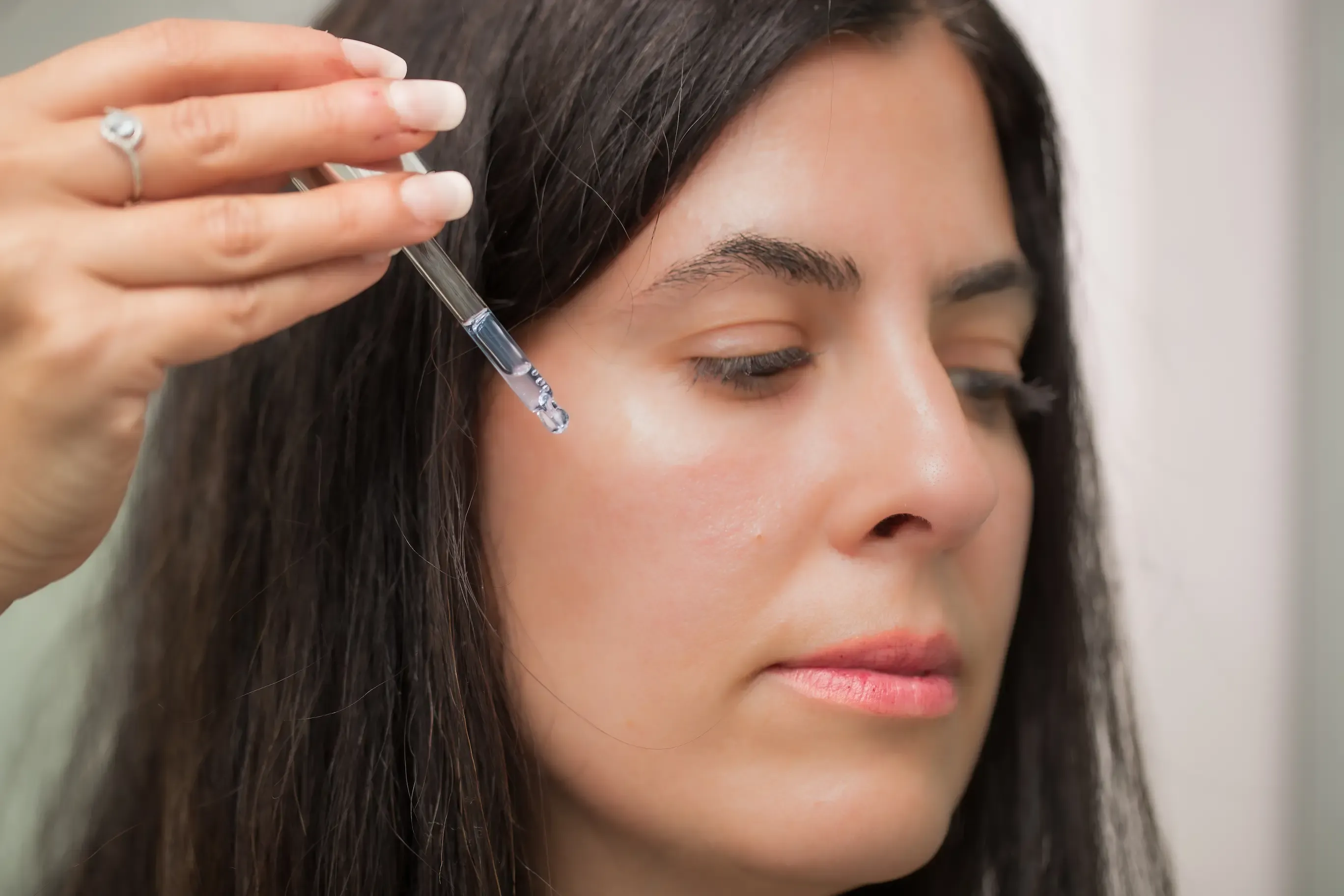 A person applying a drop of facial serum from a glass dropper onto their cheek.