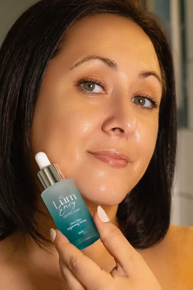 A person holds a small, teal glass bottle of facial serum near their face, which has a dewy, glowing complexion.
