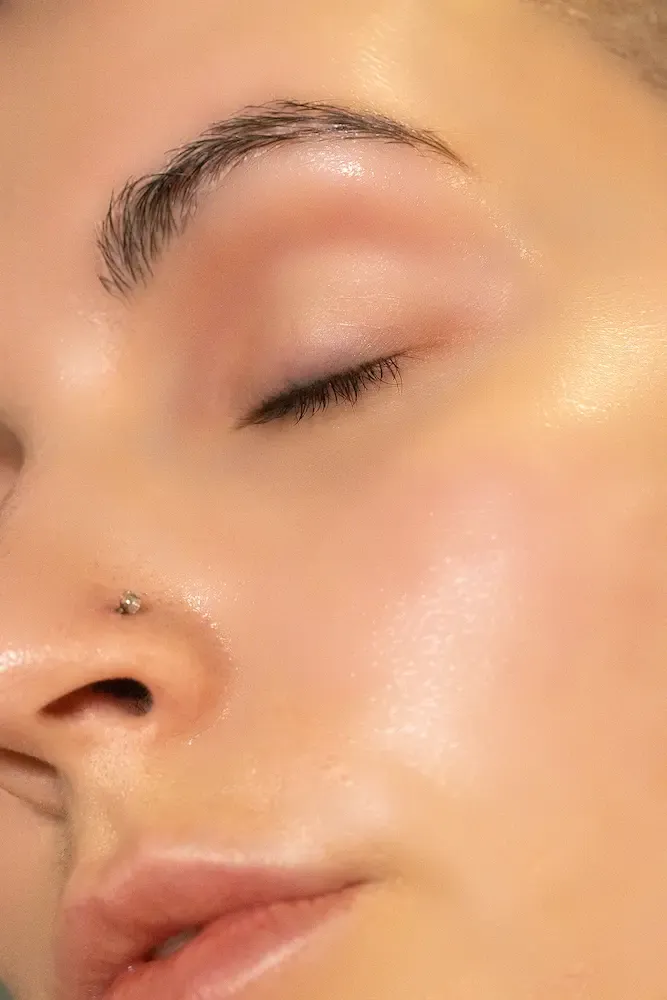 Close-up of a face with dewy, luminous skin, a small silver nose stud, and groomed eyebrows, eyes closed.