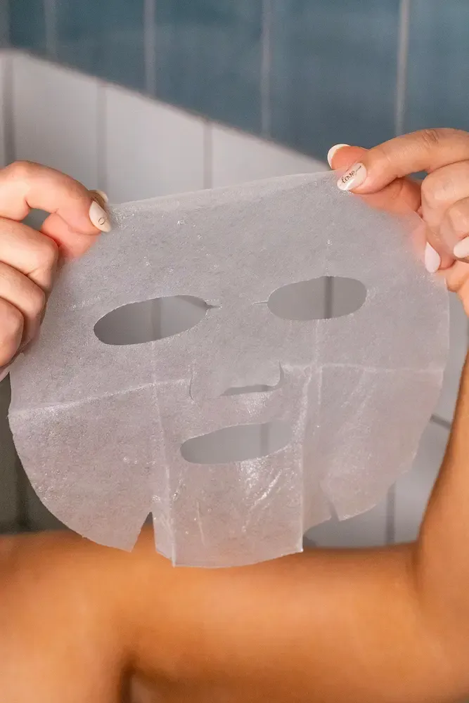 A person holds up a white, saturated sheet mask with eye, nose, and mouth cutouts against a tiled wall.