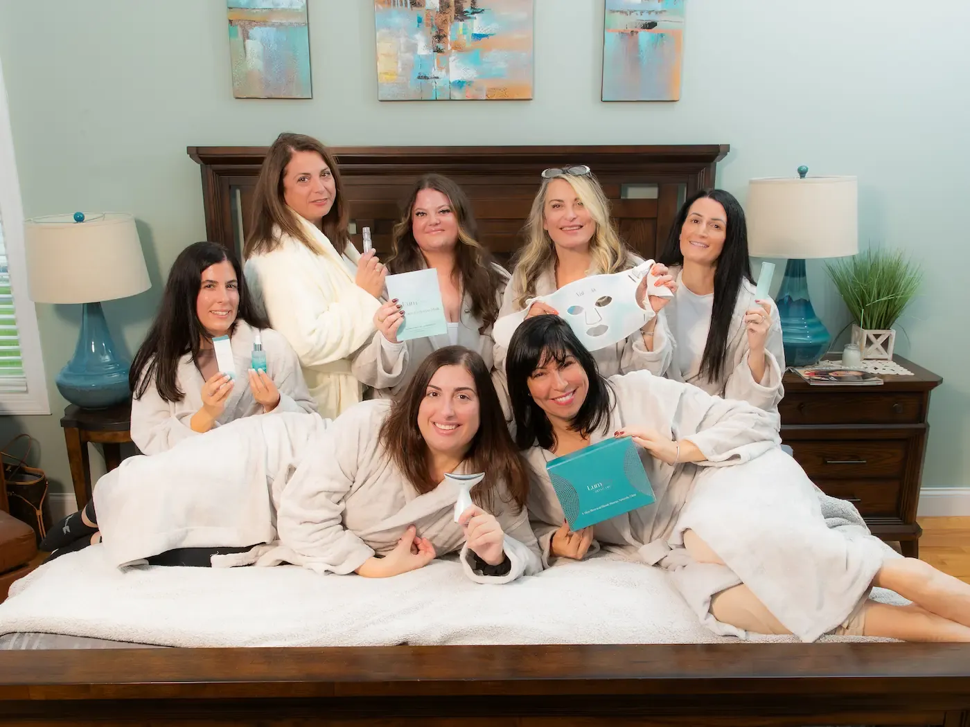 Seven smiling people in robes pose on a bed with skincare products, held against a bedroom background.