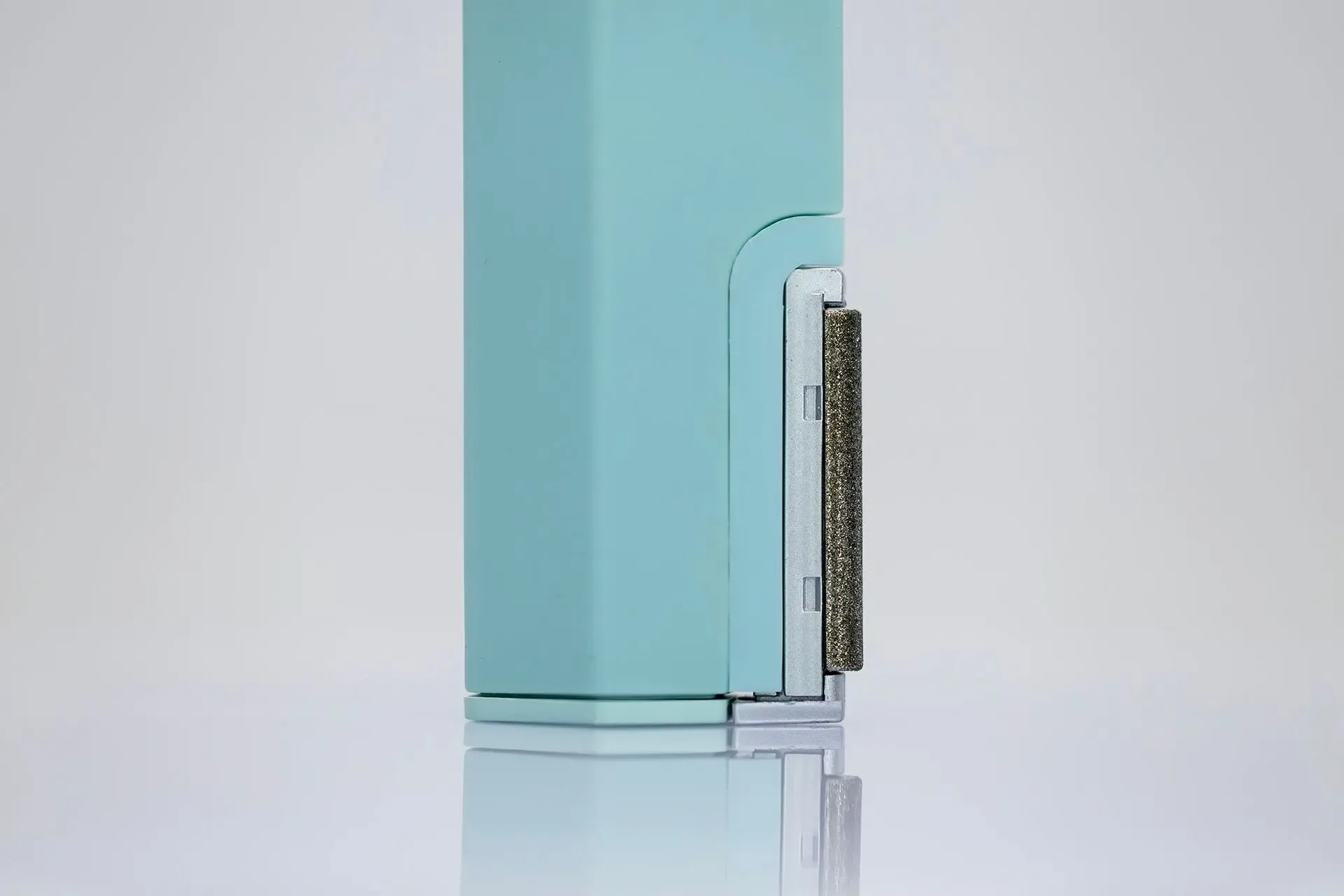 A close-up of a teal, vertical device featuring a silver rectangular attachment with a textured, grey, bristle-like side.