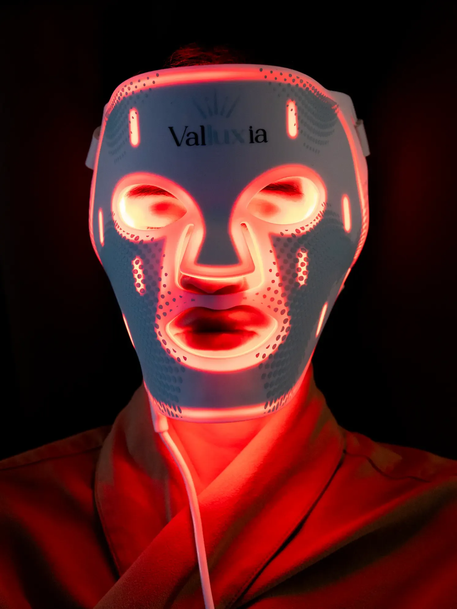 A person wears a glowing red LED light therapy mask with the brand name