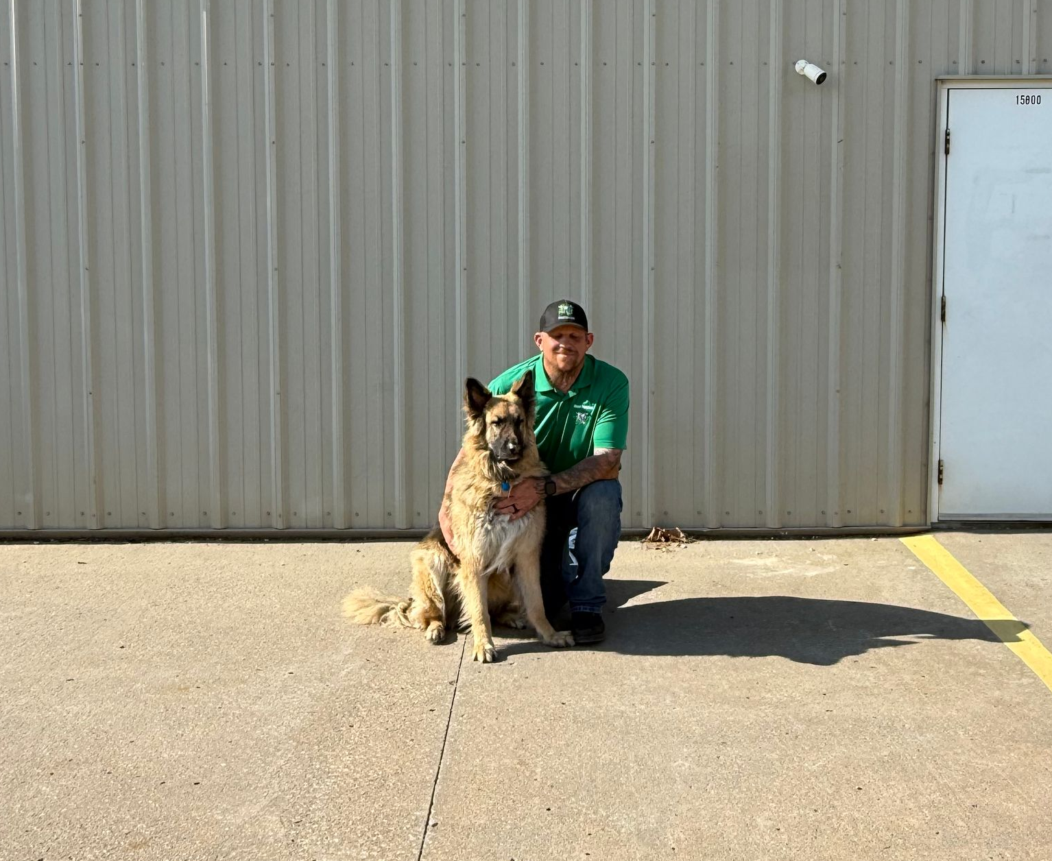 Person kneeling beside a German shepherd in front of a gray metal wall and white door.