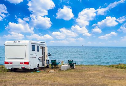 Un camper parcheggiato in riva a un lago sotto un cielo azzurro brillante con nuvole, sedie e un frigo portatile all'esterno.