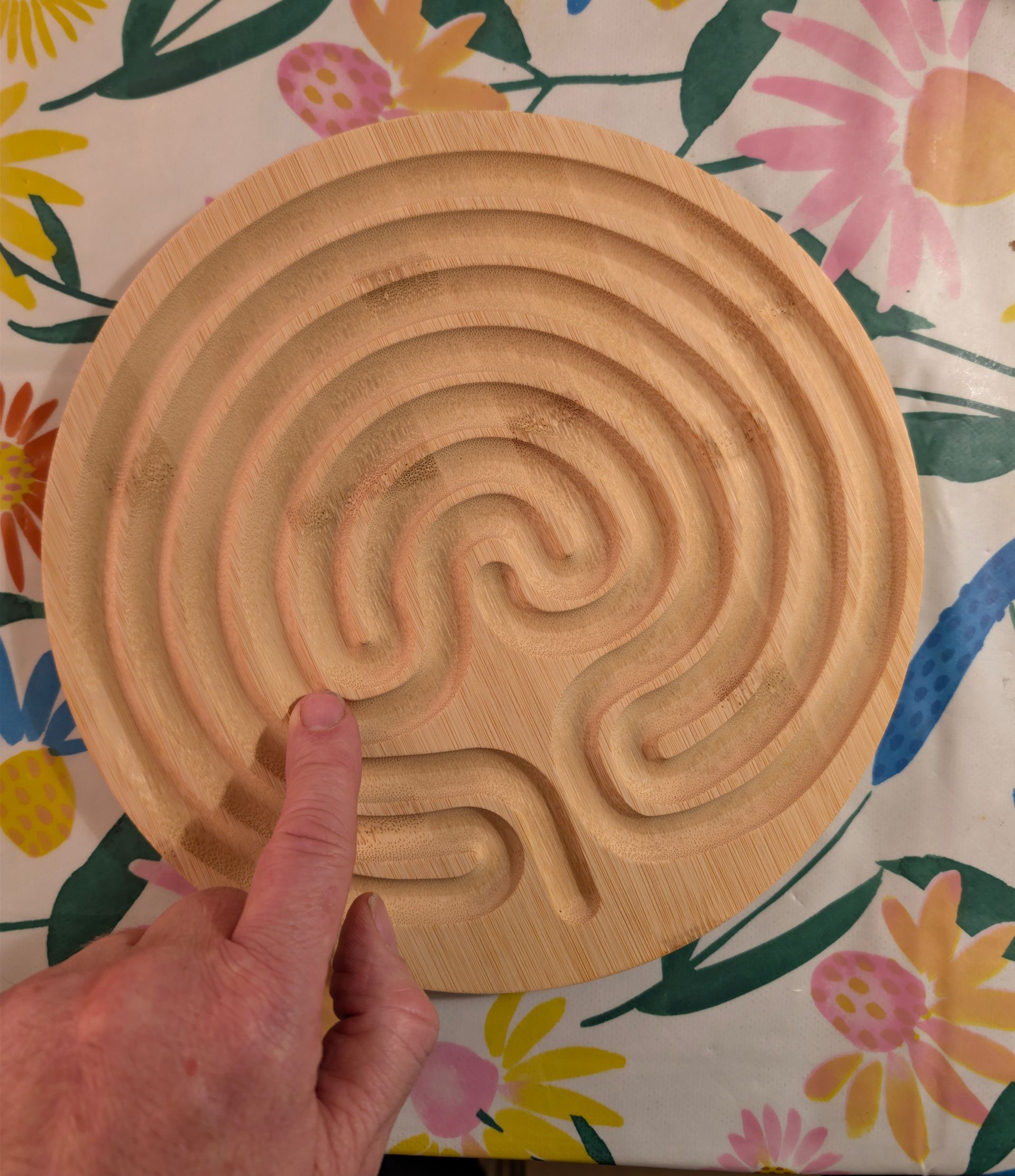 Houten labyrint met een vinger die naar het beginpad wijst. Op een tafelkleed met bloemenpatroon.