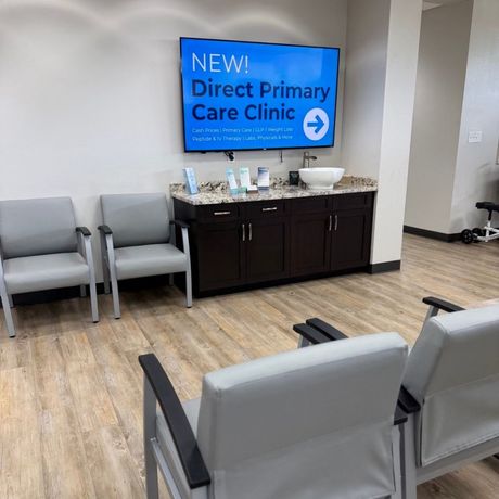 Clinic waiting room with gray chairs and a blue sign reading “NEW! Direct Primary Care Clinic”