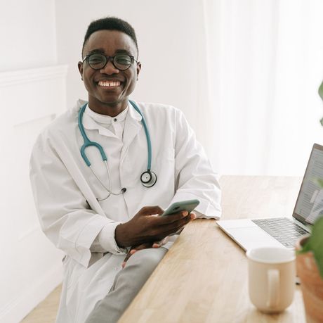 Doctor in white coat, stethoscope, and glasses smiles while holding a phone at a desk.