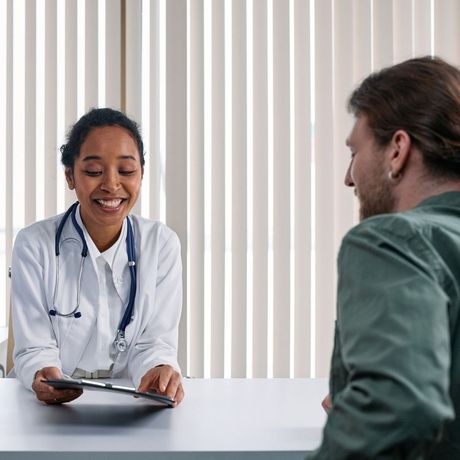 Doctor smiling, reviewing paperwork with a patient at a desk. Office setting, window blinds in background.