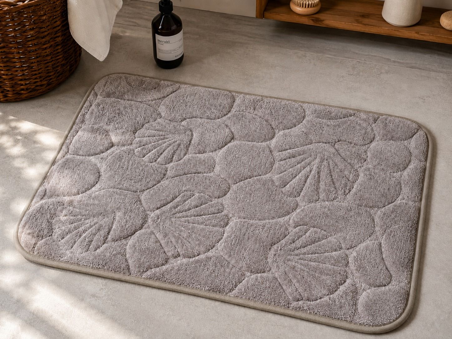 Alfombrilla de baño blanca con estampado floral sobre un suelo de madera clara, junto a una estantería y una botella.