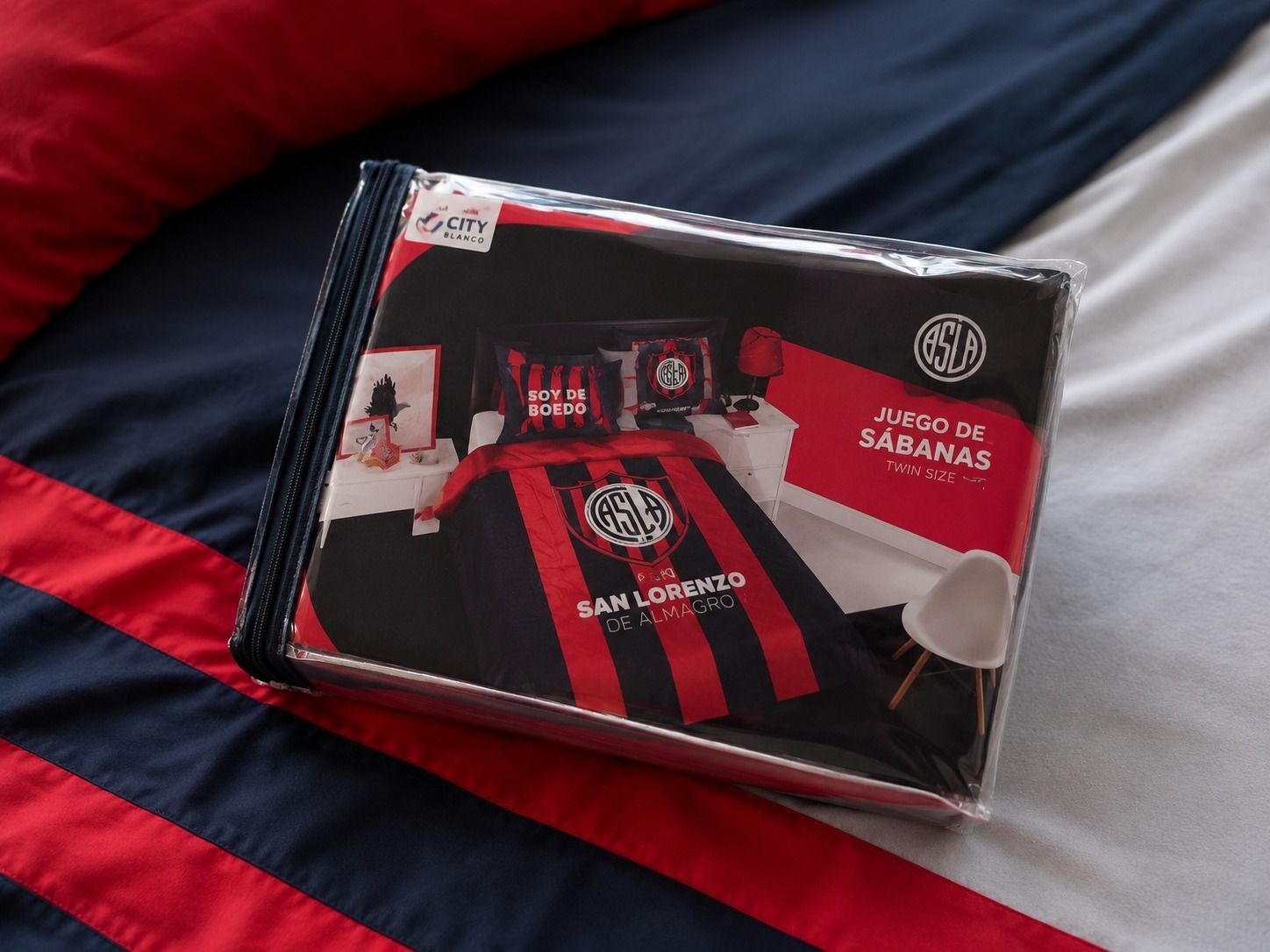 Habitación ordenada con una colcha San Lorenzo roja y azul, almohada a juego y luz cálida en la ventana.