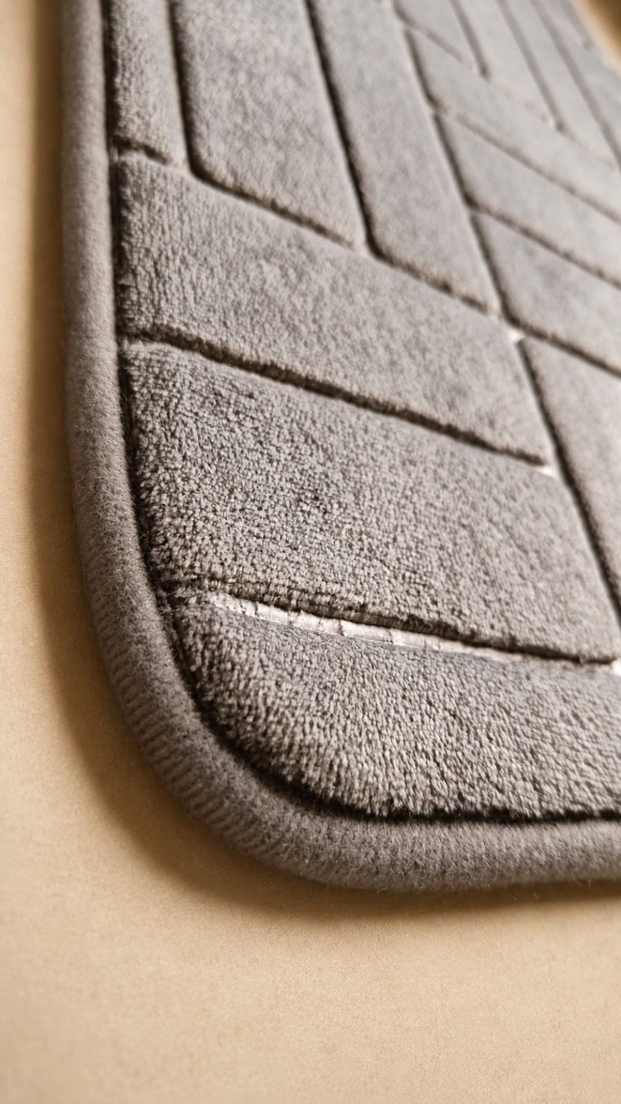 Alfombrilla de baño gris tejida sobre un suelo beige claro, junto a una estantería de madera cerca de una ventana.