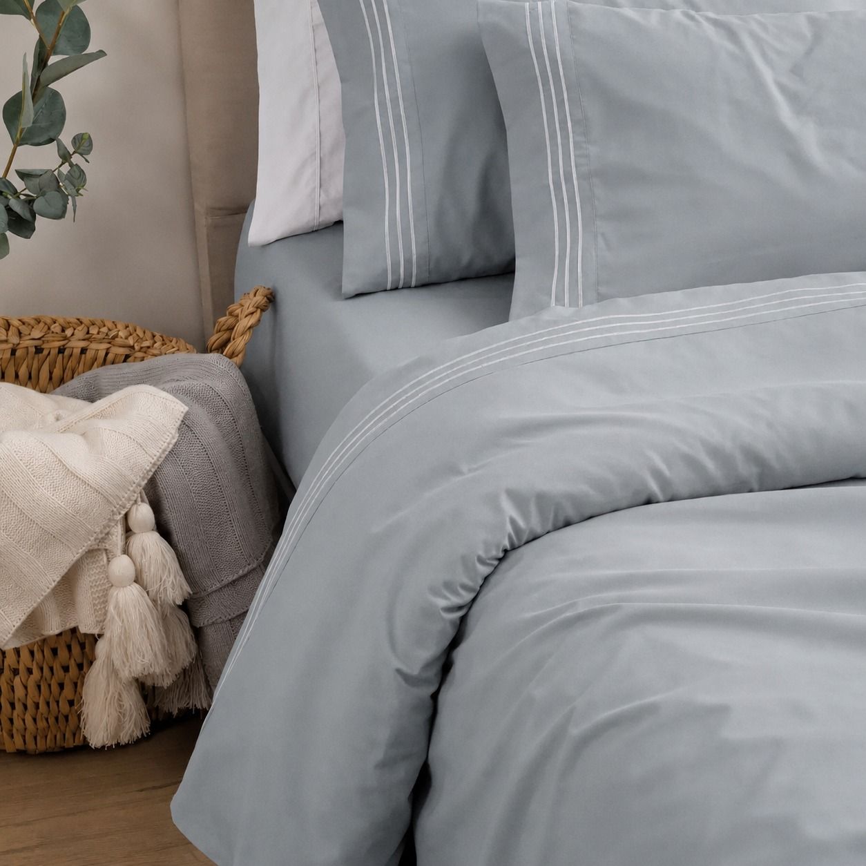 Cama bien hecha con ropa de cama gris claro, almohadas y una cesta de mimbre al lado.
