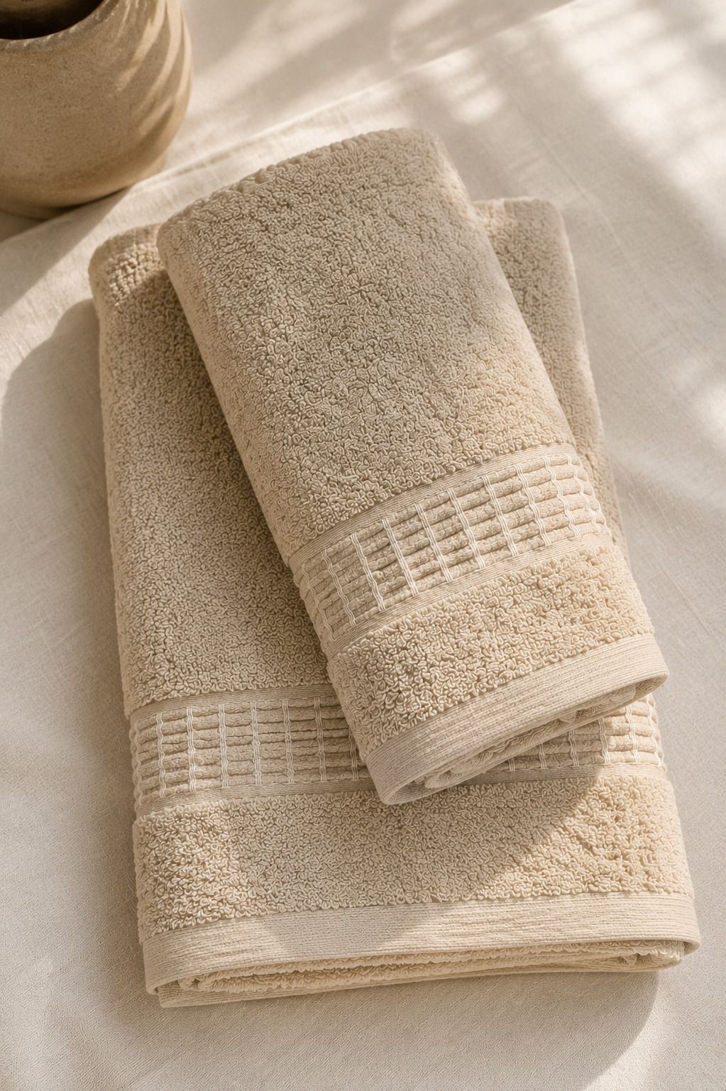 Dos toallas beige dobladas, apiladas sobre una superficie clara bajo una luz solar suave.
