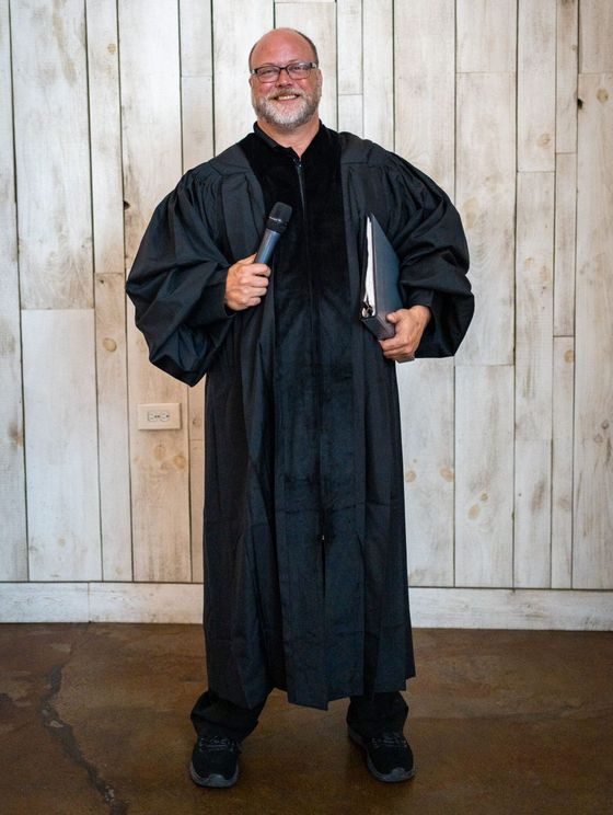 A man in a graduation gown is holding a microphone and a book.