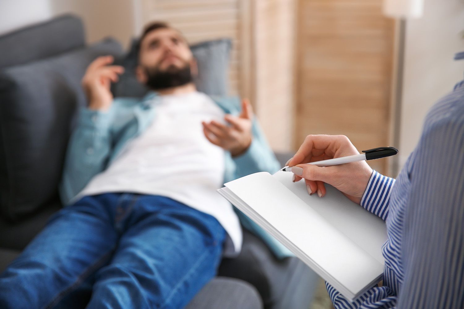 Therapist taking notes while a person lies on a couch, talking. Indoor setting.