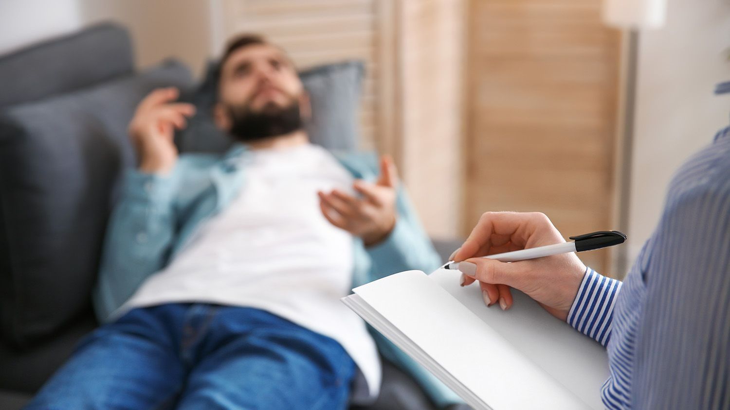 A person lies on a couch, talking to a person holding a notebook and pen.