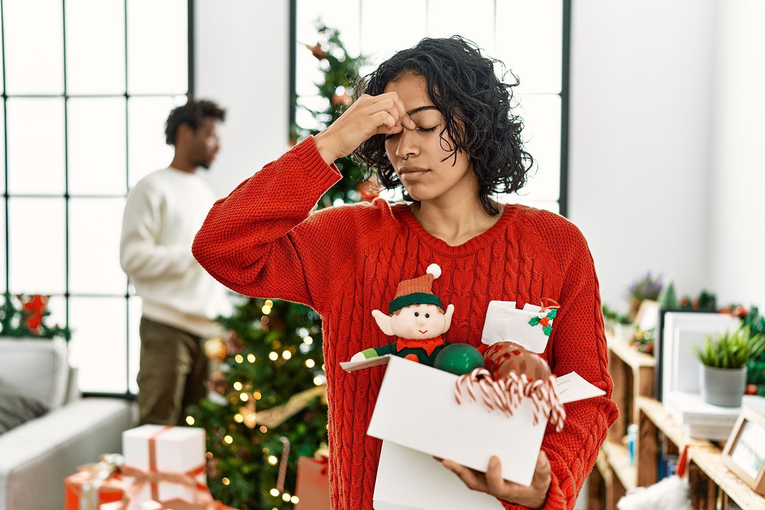 Woman holding Christmas decorations, pinching her nose, appearing stressed. Man in background near tree.