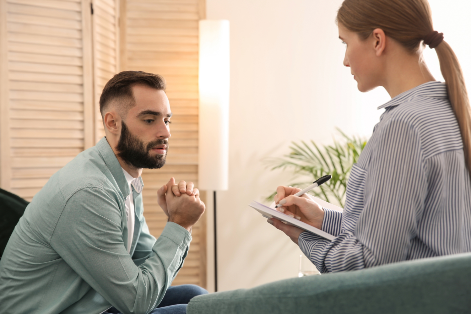 Man in therapy session, seated, hands clasped. Therapist taking notes, facing him. Softly lit room, plant in background.