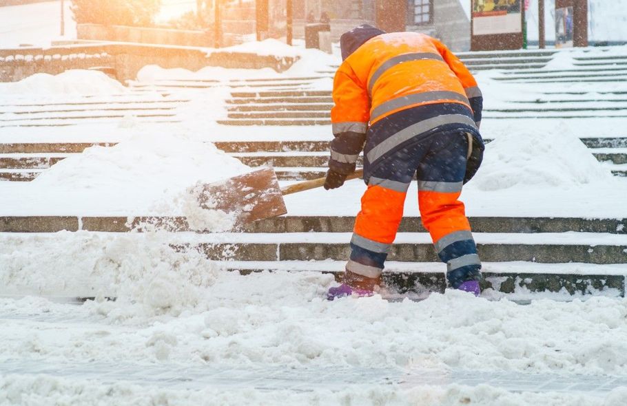 Worker in orange safety gear shoveling snow from outdoor steps.