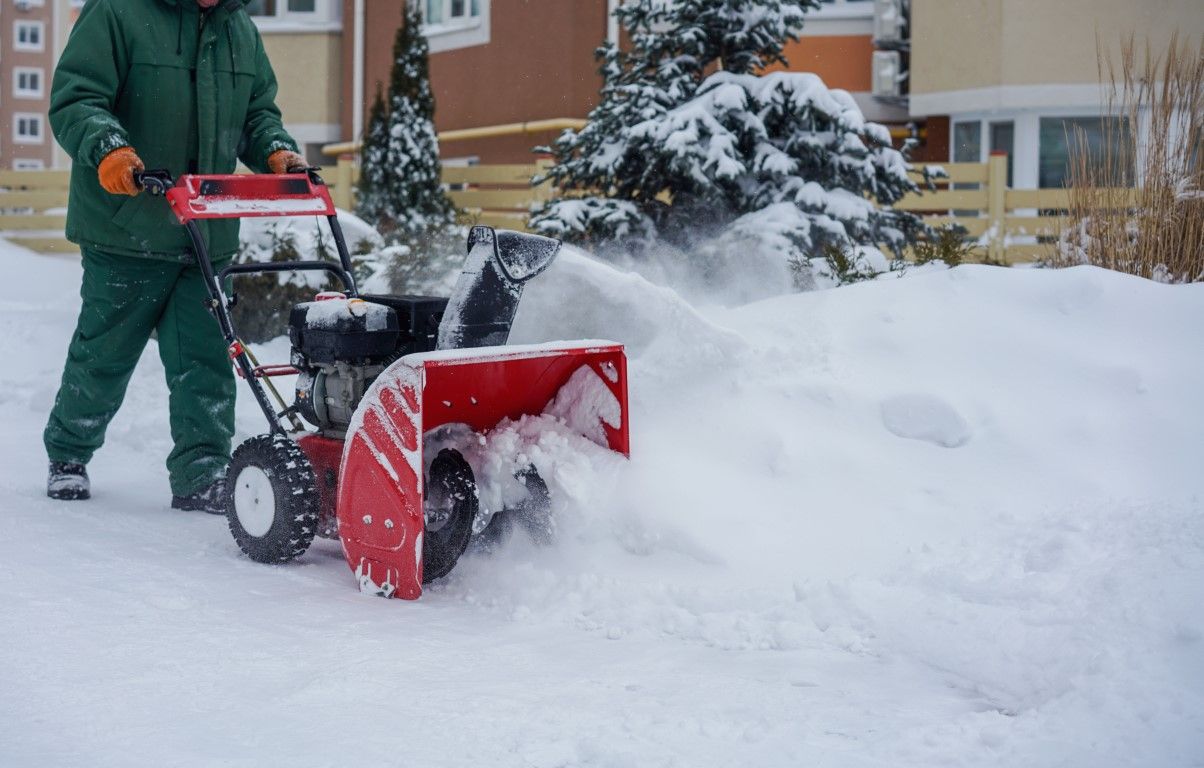 Person using a snow blower to clear a snowy driveway near apartment buildings