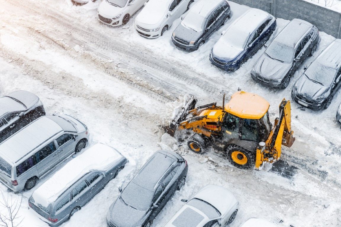 Snowy parking lot with a yellow snowplow clearing snow between parked cars.