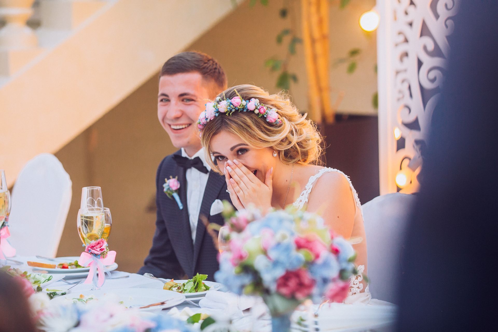 A bride in a floral crown laughs while covering her mouth, sitting next to a smiling groom at a wedding reception table. A bride in a floral crown laughs while covering her mouth, sitting next to a smiling groom at a wedding reception table.