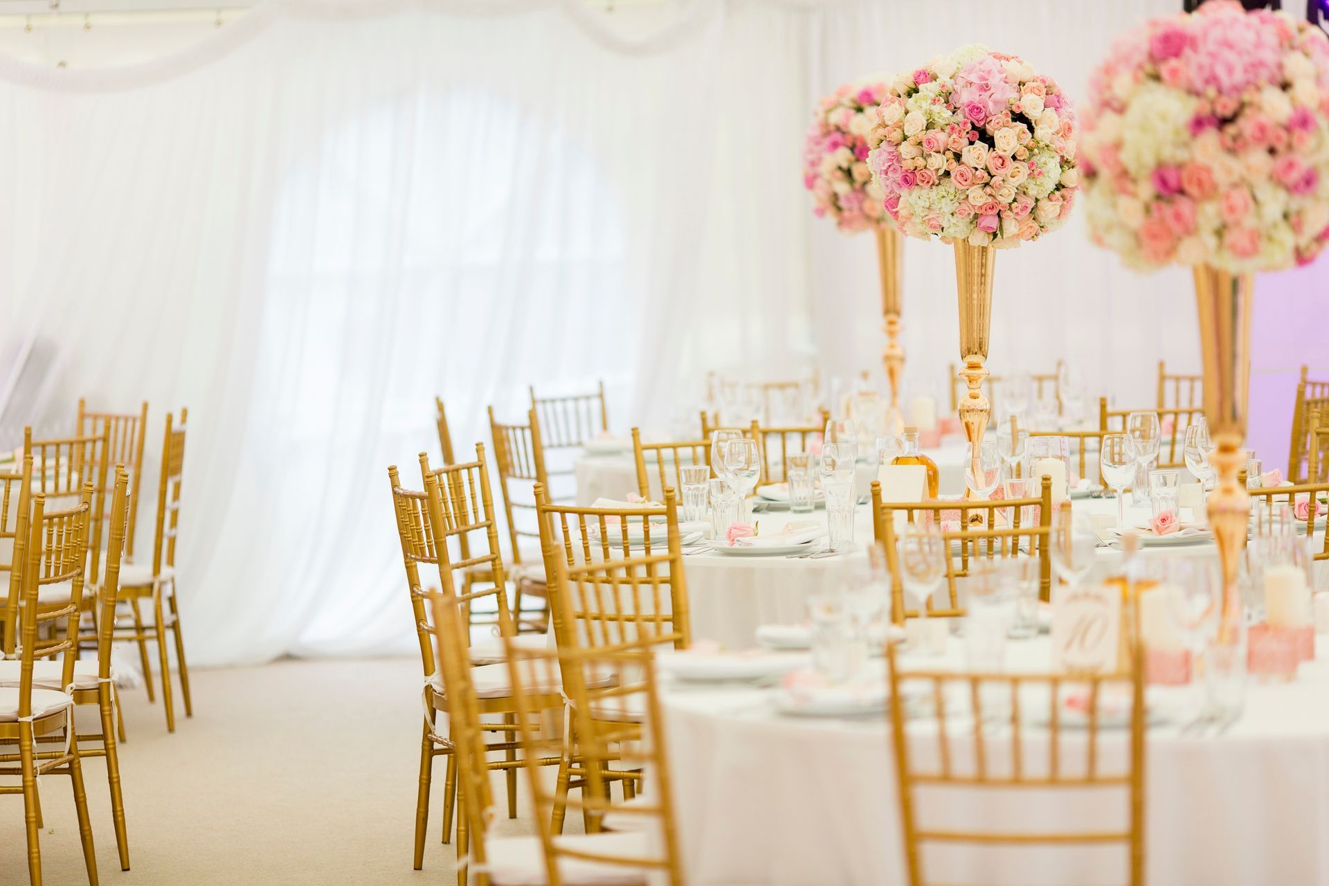 Elegant round banquet tables with gold chairs and tall floral centerpieces inside a draped white tent.