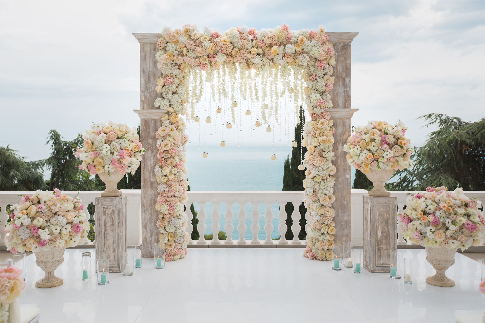A wedding altar covered in light pink and white flowers, centered on a white terrace overlooking the ocean.