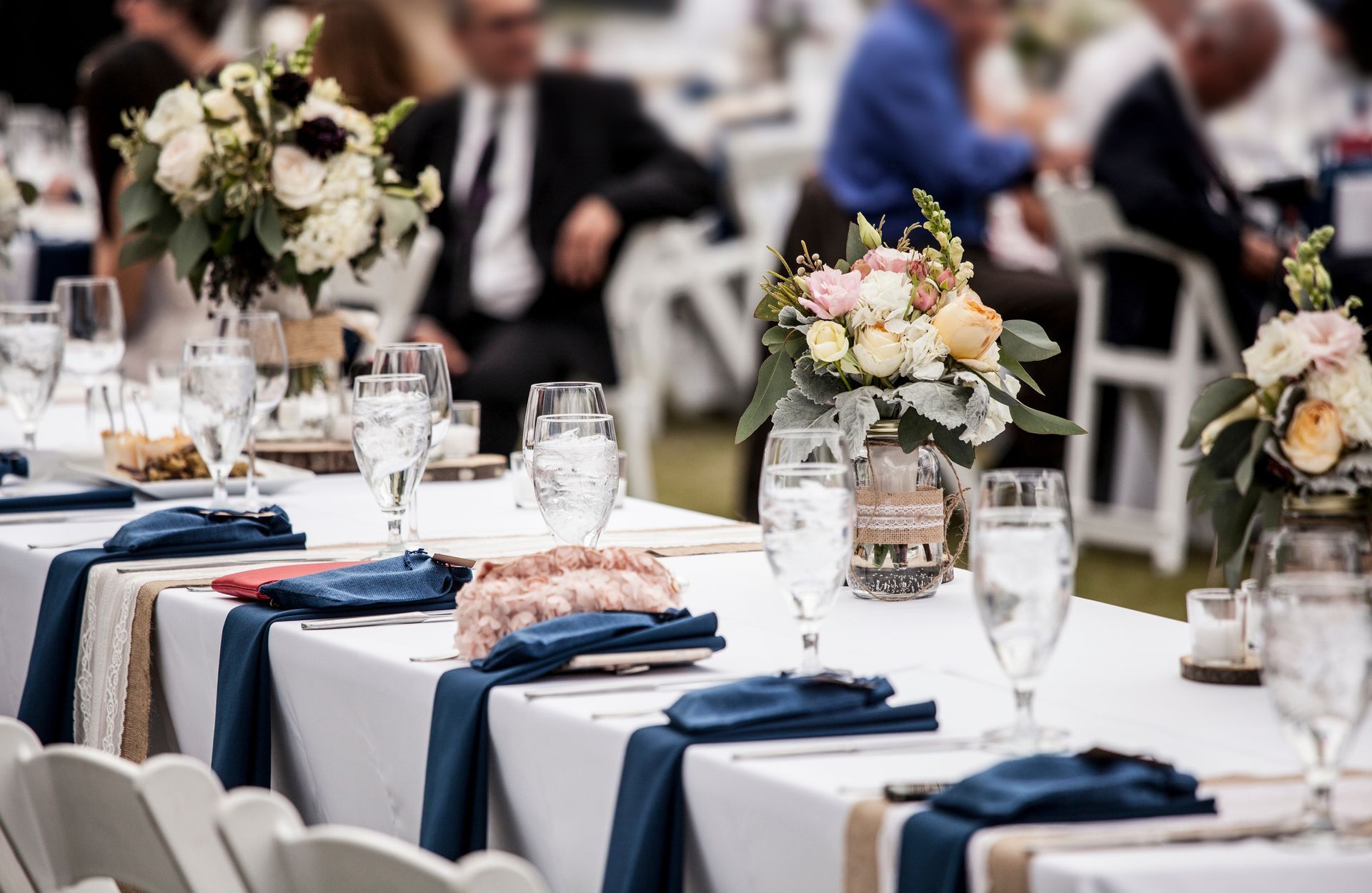 A long banquet table set with white tablecloths, navy napkins, glassware, and floral centerpieces at an event. A long banquet table set with white tablecloths, navy napkins, glassware, and floral centerpieces at an event.