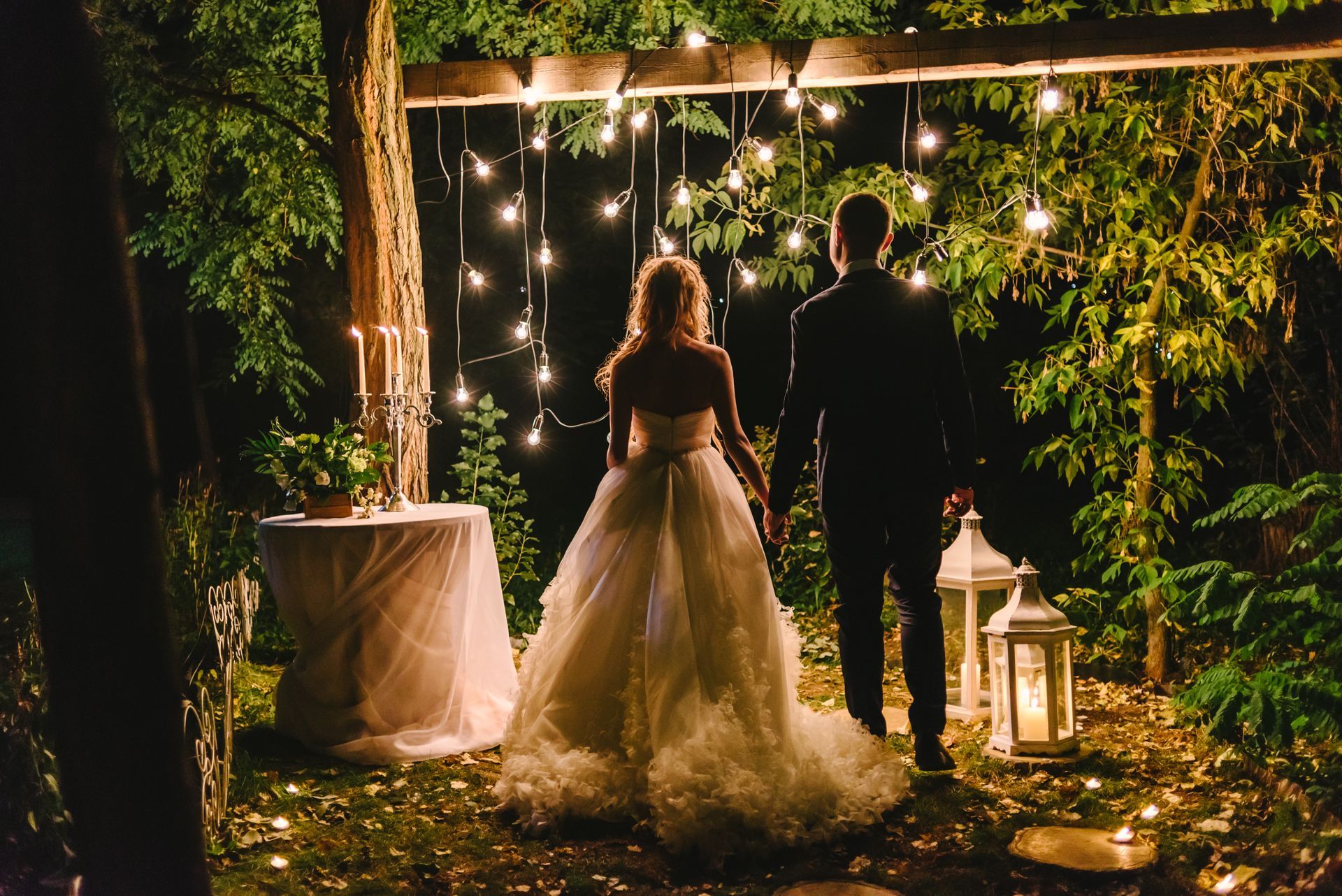 A couple standing hand-in-hand in a garden at night, illuminated by string lights and lanterns. A couple standing hand-in-hand in a garden at night, illuminated by string lights and lanterns.