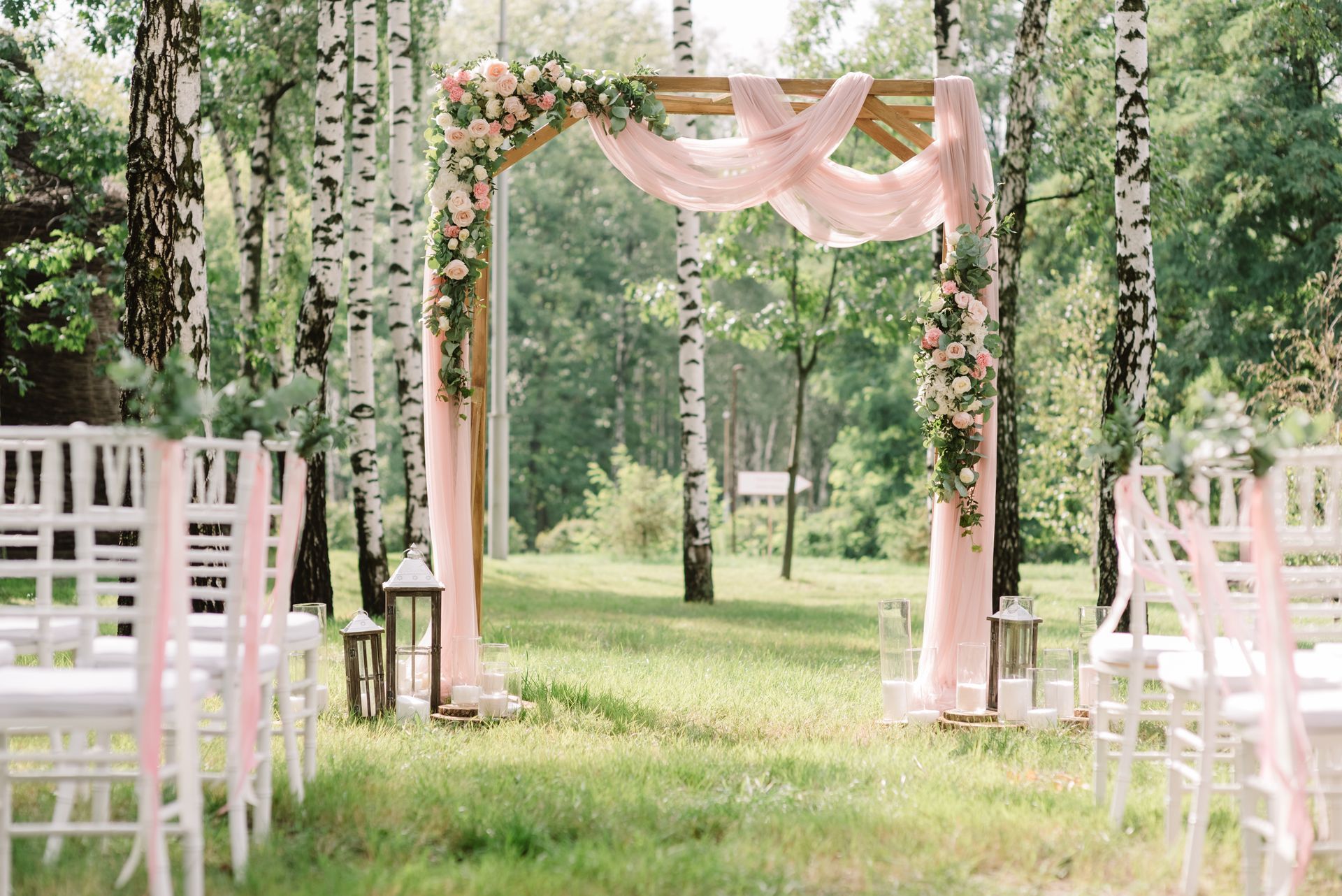 Outdoor wedding aisle with white chairs, pink drapes, and floral garlands under trees Outdoor wedding aisle with white chairs, pink drapes, and floral garlands under trees