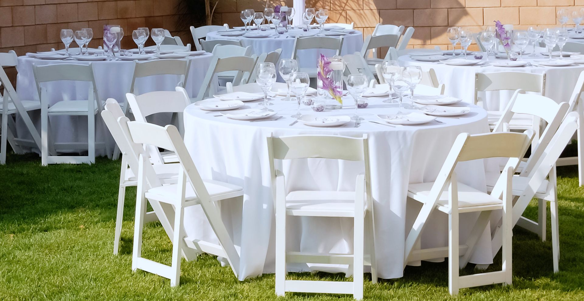 Multiple round tables with white tablecloths and folding chairs are set up on a grassy lawn for an outdoor event.
