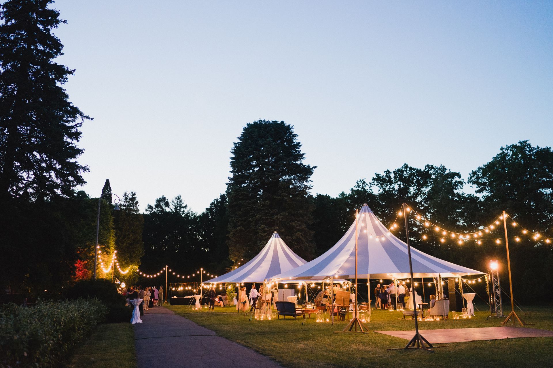 Two large white event tents illuminated by string lights at dusk in a grassy outdoor setting with trees in the background.