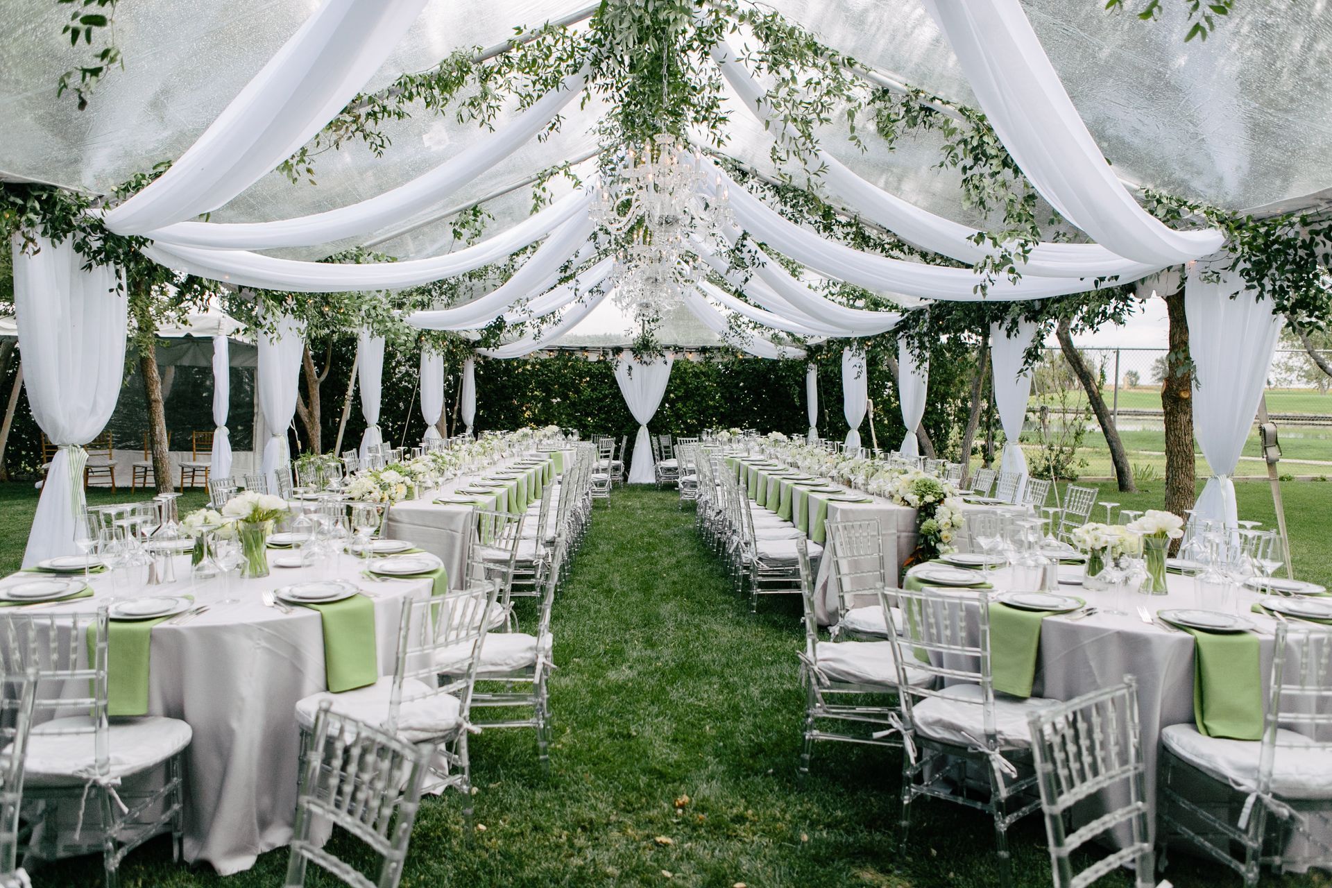 Outdoor wedding reception with draped white fabric and greenery overhead, featuring round tables with white linens.