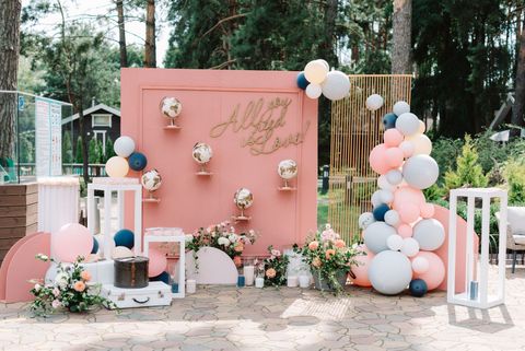A pink wedding backdrop with globes, floral arrangements, and a balloon arch, set outdoors in a wooded area.