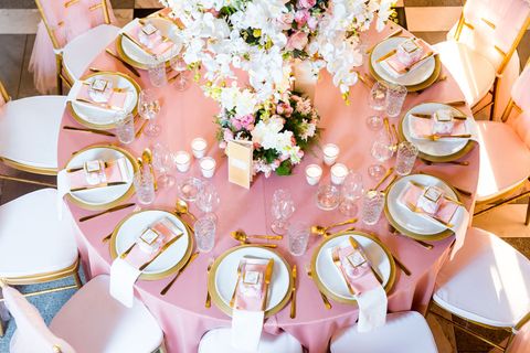 A round table with a pink tablecloth and gold-rimmed white plates, surrounded by pink chairs and centered with flowers.